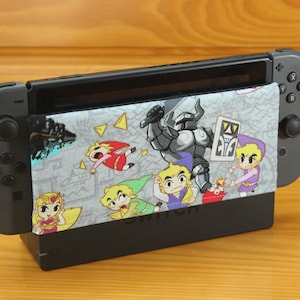 May include: A black Nintendo Switch console with a gray and white fabric cover featuring cartoon characters. The cover has a design of a knight, a sword, and several characters from the Legend of Zelda video game series. The console is sitting on a wooden surface.