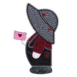 May include: A gray and black patchwork doll with a red heart on a pink envelope. The doll is wearing a burgundy shirt and black overalls. The doll is holding the envelope in its right hand.