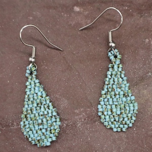 May include: A pair of beaded earrings with a teardrop shape. The earrings are made of small, light blue and green beads, and have silver-colored ear wires. The earrings are displayed on a brown surface.