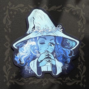 May include: A die-cut sticker featuring a stylised illustration of a figure with long wavy blue hair wearing a wide-brimmed pointed hat adorned with a white vine-like pattern. The figure has a light blue complexion, winks one eye, and holds hands together near the face, rendered in shades of dark blue, light blue, and white.