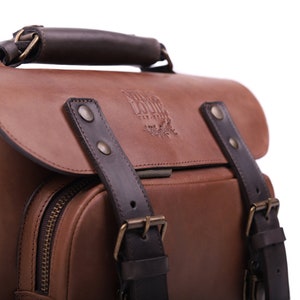May include: A brown leather satchel with a dark brown leather handle and straps. The bag has a zippered pocket on the front and a brass buckle on the strap. The bag has a logo embossed on the front that reads 'Wild Bound'.
