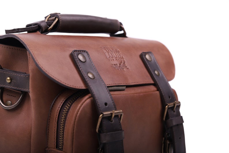 May include: A brown leather satchel with a dark brown leather handle and straps. The bag has a zippered pocket on the front and a brass buckle on the strap. The bag has a logo embossed on the front that reads 'Wild Bound'.