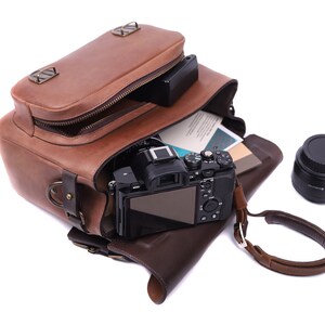 May include: A brown leather camera bag with a camera and lens inside. The bag is open and the camera is visible. The camera is black and has a silver lens. The lens is also black and has a silver ring around it. The bag has a brown leather strap and a metal buckle.