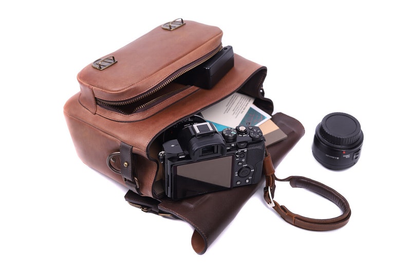 May include: A brown leather camera bag with a camera and lens inside. The bag is open and the camera is visible. The camera is black and has a silver lens. The lens is also black and has a silver ring around it. The bag has a brown leather strap and a metal buckle.