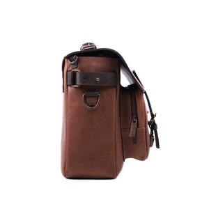 May include: A brown leather messenger bag with a black strap and buckle. The bag has a front pocket with a zipper and a side pocket with a buckle. The bag is on a white background.