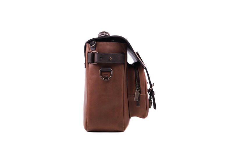 May include: A brown leather messenger bag with a black strap and buckle. The bag has a front pocket with a zipper and a side pocket with a buckle. The bag is on a white background.
