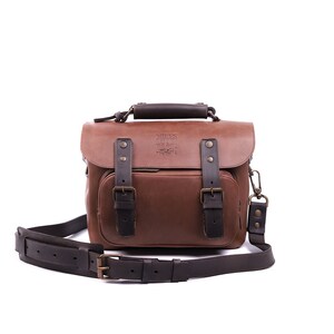 May include: A brown leather camera bag with a black strap and buckles. The bag has a logo that says 'James Henry Co. Handmade.' 