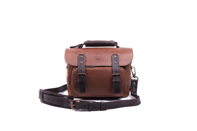 May include: A brown leather camera bag with a black strap and buckles. The bag has a logo that says 'James Henry Co. Handmade.' 