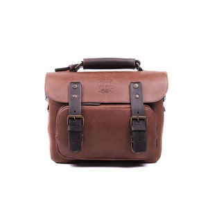 May include: A brown leather camera bag with black straps and buckles. The bag has a top handle and a front flap with a brass buckle. The bag has a logo that reads 'Atlas Photo Co.' on the front flap.