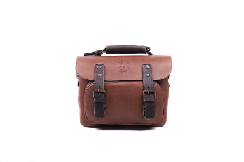 May include: A brown leather camera bag with black straps and buckles. The bag has a top handle and a front flap with a brass buckle. The bag has a logo that reads 'Atlas Photo Co.' on the front flap.
