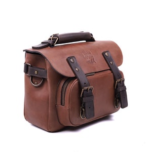 May include: A brown leather camera bag with a dark brown leather handle and buckles. The bag has a zippered pocket on the front and a logo that reads 'Nomad Goods'.