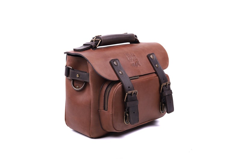 May include: A brown leather camera bag with a dark brown leather handle and buckles. The bag has a zippered pocket on the front and a logo that reads 'Nomad Goods'.