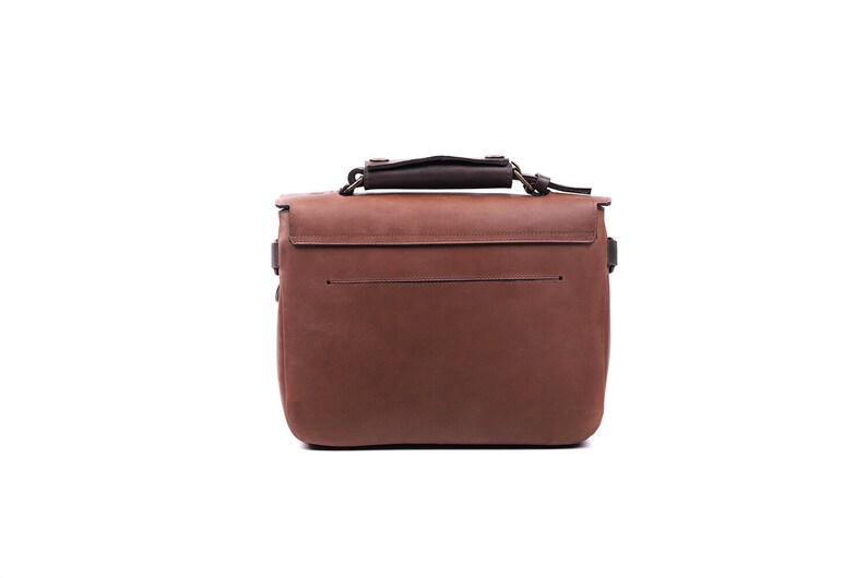 May include: A brown leather satchel bag with a top handle and a flap closure. The bag has a front pocket and a back pocket. The bag is made of high-quality leather and has a classic design.