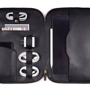 Puede incluir: Funda para portátil de cuero negro con cierre de cremallera. La funda tiene varios compartimentos para organizar cables, un teléfono y un lápiz. Se muestra una tableta dentro de la funda.