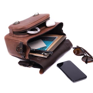 May include: A brown leather satchel bag open with a white phone, a notebook, pens, and sunglasses inside. The bag has a zipper and a buckle closure.