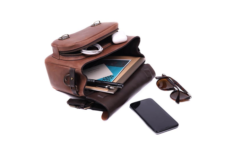 May include: A brown leather satchel bag open with a white phone, a notebook, pens, and sunglasses inside. The bag has a zipper and a buckle closure.