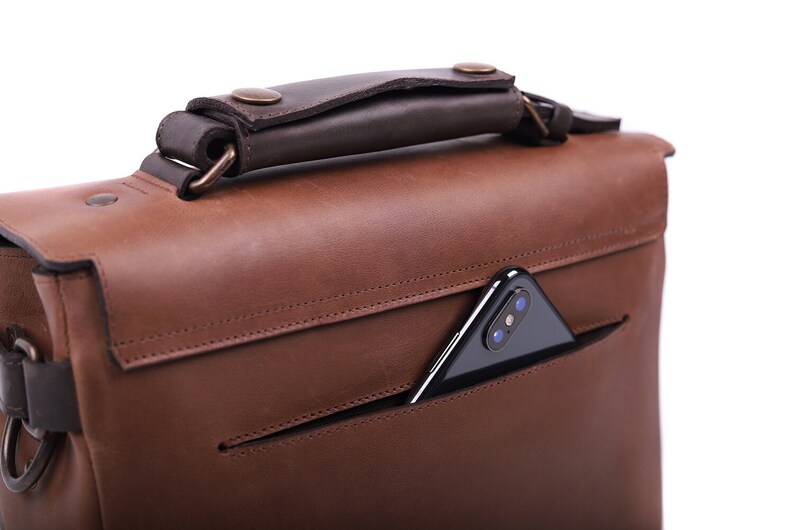 May include: A brown leather messenger bag with a dark brown leather handle. The bag has a pocket on the front with a black smartphone inside.