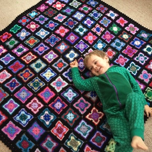 May include: A child wearing a green and black patterned onesie lies on a black and multi-colored crocheted blanket. The blanket is made of squares with different colored yarn.