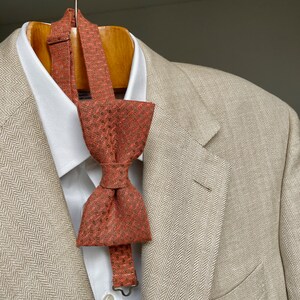 May include: A beige herringbone blazer with a brown patterned bow tie. The bow tie is tied and hangs down the front of the blazer.