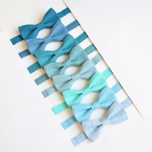 May include: A set of seven bow ties in shades of blue and teal, arranged in a gradient from dark blue to light teal. The bow ties are made of a soft, textured fabric.