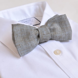 Puede incluir: Una pajarita gris de tela texturizada descansa sobre una camisa blanca. La pajarita está centrada en el cuello de la camisa, con el botón superior abrochado. La pajarita tiene una forma clásica y un color neutro.