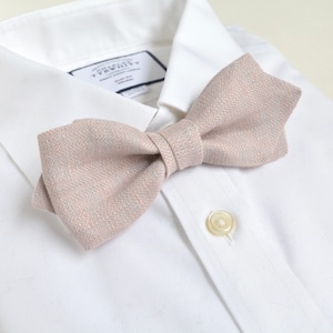 Può includere: Un papillon rosa chiaro con un motivo strutturato è appoggiato su una camicia bianca. Il papillon è annodato al centro e ha una forma classica. La camicia ha un colletto button-down e una chiusura con un solo bottone.