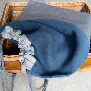 Può includere: Una borsa blu con coulisse e un accento a righe è in un cestino di legno. Un tessuto grigio è piegato nel cestino dietro la borsa. La borsa ha una chiusura con coulisse e un cordino abbinato. Il cestino è in legno intrecciato.
