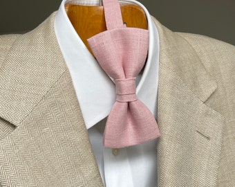 May include: A light pink bow tie made of textured fabric, displayed on a white collared shirt and a tan herringbone blazer. The bow tie is knotted and ready to wear, perfect for formal occasions.