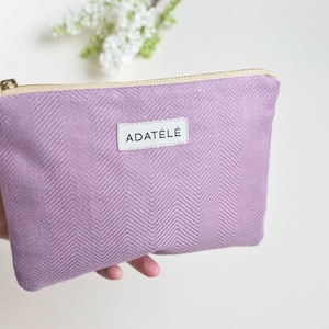 Può includere: Una pochette con cerniera color lavanda con un motivo a spina di pesce e una cerniera dorata. La pochette ha una piccola etichetta bianca con il testo "ADATÉLÉ" in nero.