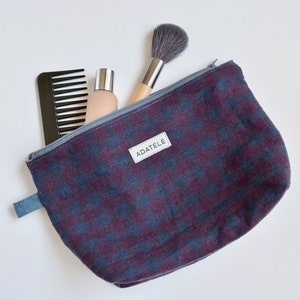 Può includere: Una trousse da trucco in tessuto a quadri blu e bordeaux con chiusura a zip e tiretto grigio. La trousse ha un'etichetta bianca con il testo "ADATÉLÉ". La trousse è piena di un pettine nero, una bottiglia di lozione e un pennello da trucco.