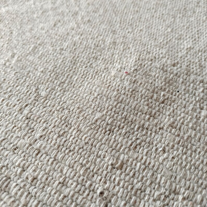 May include: Close-up of a textured, off-white woven fabric. The weave is tight, creating a slightly ridged surface. The fabric appears to be made of natural fibers, with subtle variations in color and texture. The overall look is neutral and organic.