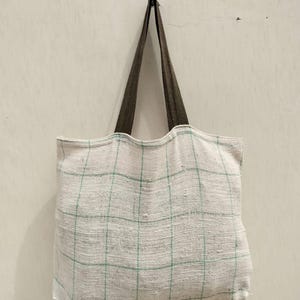 May include: A white tote bag with a green grid pattern hangs from a black hook. The bag has brown straps and is made of a textured fabric. The bag is empty and hangs against a plain white wall.