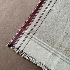 May include: Close-up of a vintage, rectangular rug with a cream-colored background. The rug features vertical stripes in a deep burgundy color, with thinner stripes in between. The edge of the rug has a fringe.