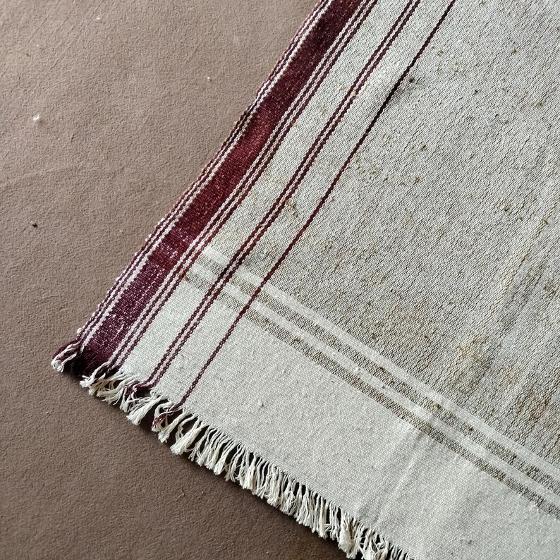 May include: Close-up of a vintage, rectangular rug with a cream-colored background. The rug features vertical stripes in a deep burgundy color, with thinner stripes in between. The edge of the rug has a fringe.