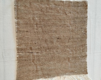 May include: A rectangular, woven textile in shades of brown with white fringe along two edges. The fabric has a textured appearance, with visible threads creating a slightly uneven surface. The fringe is made of white, unraveled threads.