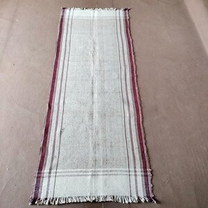 May include: Cream-colored rectangular rug with burgundy stripes along the edges and a fringed end. The rug has a simple, rustic design, suitable for a hallway or entryway. The rug is made of a woven material.