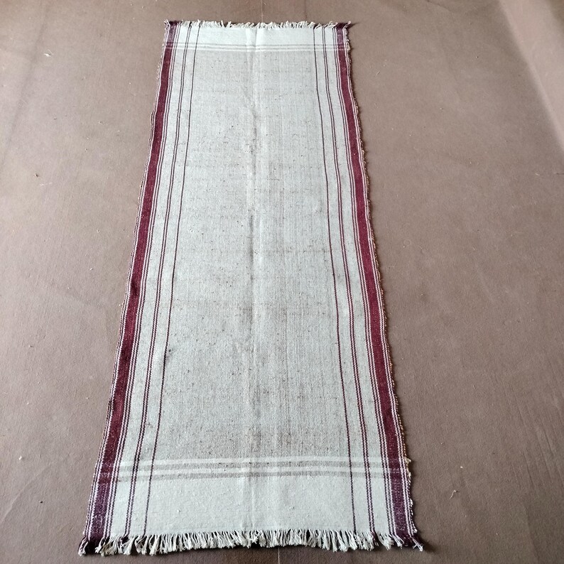 May include: Cream-colored rectangular rug with burgundy stripes along the edges and a fringed end. The rug has a simple, rustic design, suitable for a hallway or entryway. The rug is made of a woven material.
