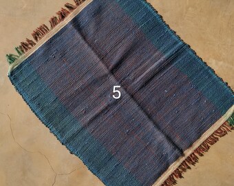 May include: A square, woven placemat with a blue and brown color scheme. The placemat has a fringed edge with green and brown accents. The number "5" is printed in white in the center of the placemat.