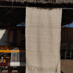 May include: A beige woven textile with a fringed edge, suspended from a metal bar. The textile is displayed outside, beneath a thatched roof. A loom is visible in the background. The textile appears to be hand-woven, possibly a rug or wall hanging.