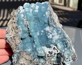 May include: A large, blue and white mineral specimen with a rough, textured surface. The mineral has a bubbly, porous texture and is set against a grey background.