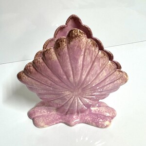 May include: A ceramic seashell-shaped decorative piece in shades of pink and brown. The fan-like structure has a textured surface with a scalloped edge. The base is also pink and has a wavy shape.