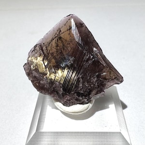May include: A faceted, dark purple mineral specimen with golden, reflective inclusions. The crystal is mounted on a clear acrylic stand, showcasing its natural, geometric form and color variations. The mineral's surface has a textured appearance.