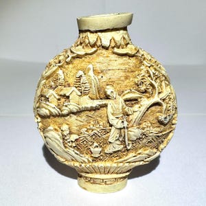 May include: A cream-colored, round snuff bottle featuring intricate, raised relief carvings of a landscape scene. The design depicts a figure standing near a small house, trees, and textured ground patterns, with a decorative base and a narrow neck.