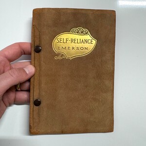 K&ouml;nnte beinhalten: Ein braunes, ledergebundenes Buch mit goldenen Lettern auf dem Einband. Der Titel lautet "Self-Reliance" von Emerson.
