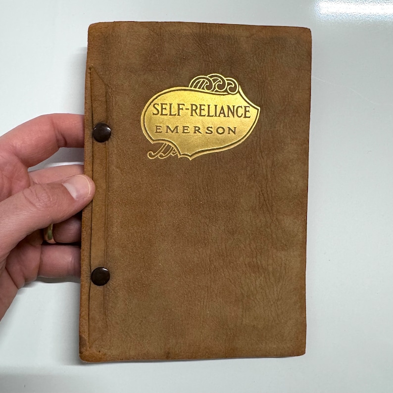 K&ouml;nnte beinhalten: Ein braunes, ledergebundenes Buch mit goldenen Lettern auf dem Einband. Der Titel lautet "Self-Reliance" von Emerson.