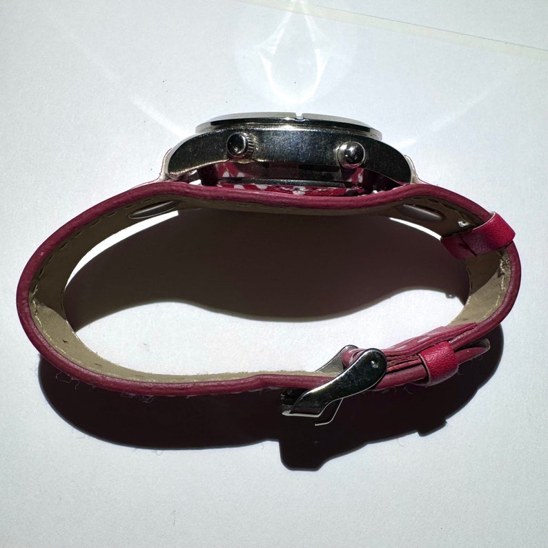 May include: A silver wristwatch with a red leather band. The band has a white polka dot pattern. The watch has a silver buckle.