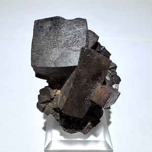 May include: A dark, metallic mineral specimen with a cluster of cubic and rectangular crystal formations. The crystals have a dark gray color and a slightly reflective surface. The mineral is displayed on a clear acrylic stand.