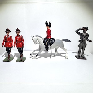 May include: A collection of vintage toy soldiers. Two soldiers in red jackets and hats stand next to a soldier on a white horse. A fourth soldier in a dark uniform salutes. The toys are on green and black bases.