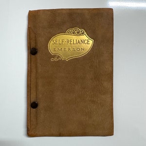 K&ouml;nnte beinhalten: Ein braunes, ledergebundenes Buch mit goldenen Lettern auf dem Einband. Der Titel lautet "Self-Reliance" von Emerson.