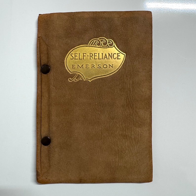 K&ouml;nnte beinhalten: Ein braunes, ledergebundenes Buch mit goldenen Lettern auf dem Einband. Der Titel lautet "Self-Reliance" von Emerson.